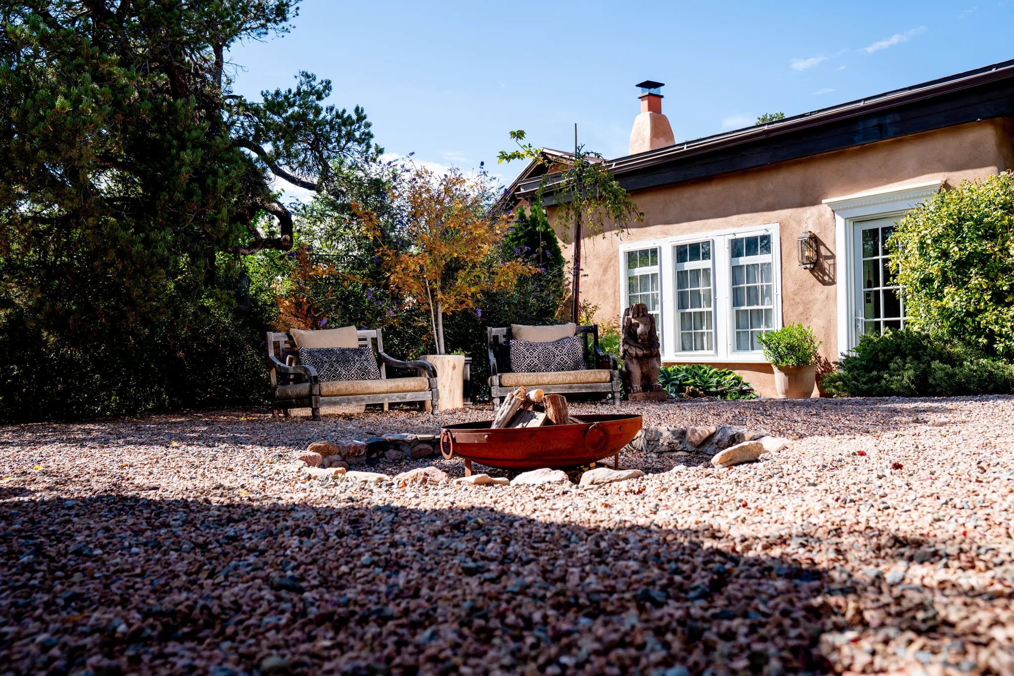 A copper firepit and two chairs on gravel beside the Los Santos adobe in afternoon light