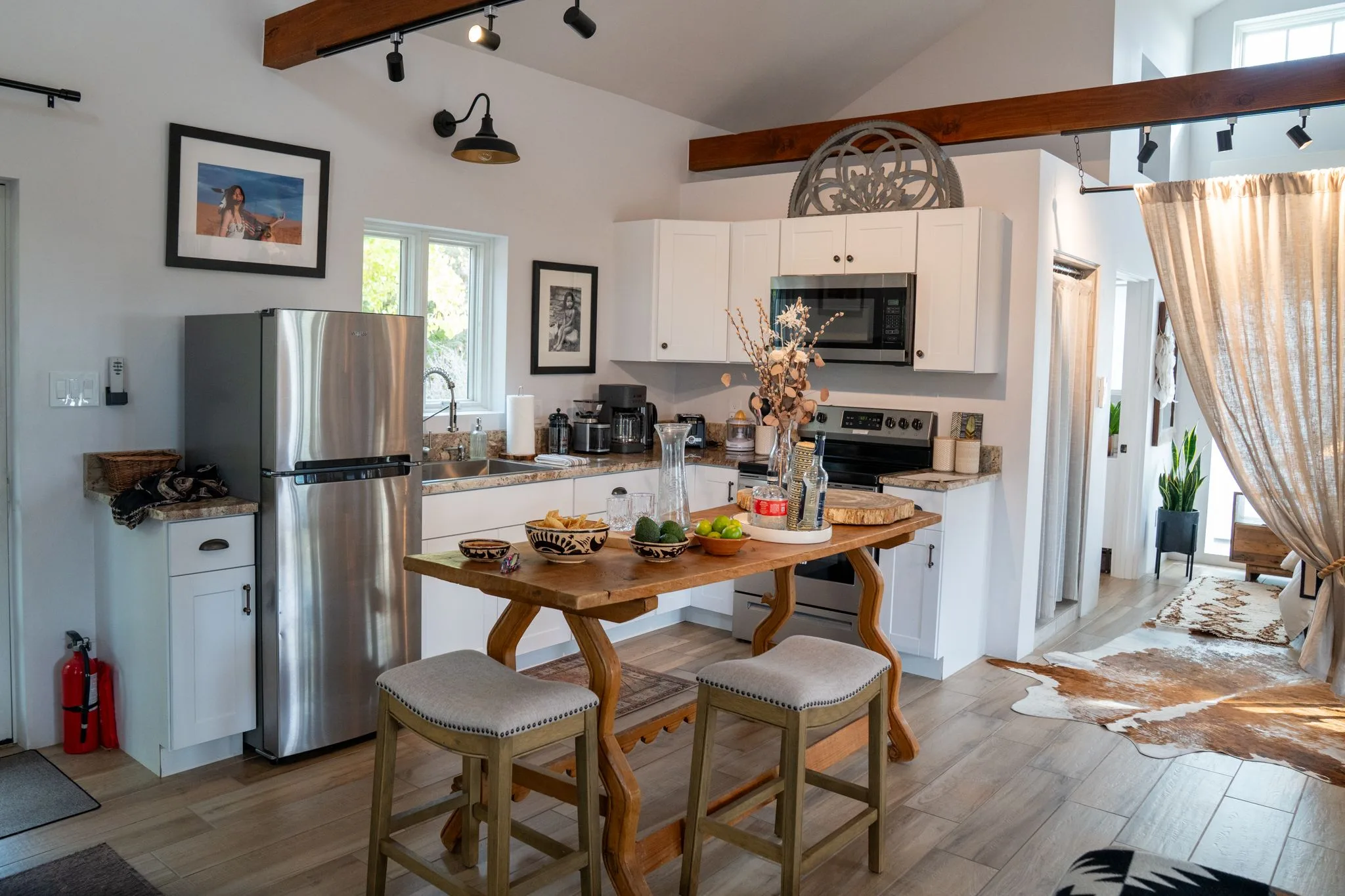 An open, light-filled room with a white kitchen, a wood island with stools, and a curtained bedroom beyond