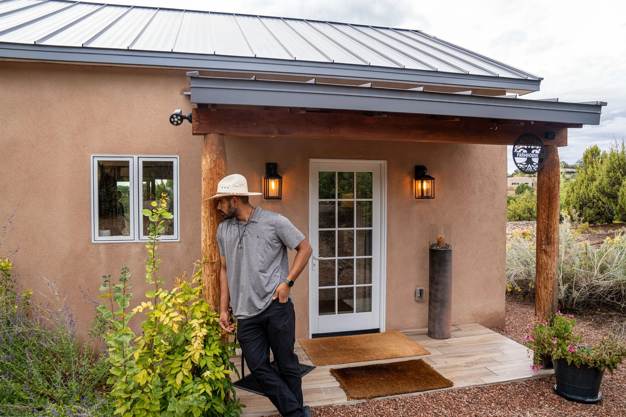 A man in a white straw hat leans by the front door of Farmhouse 1, lanterns lit, sage and flowers around the step