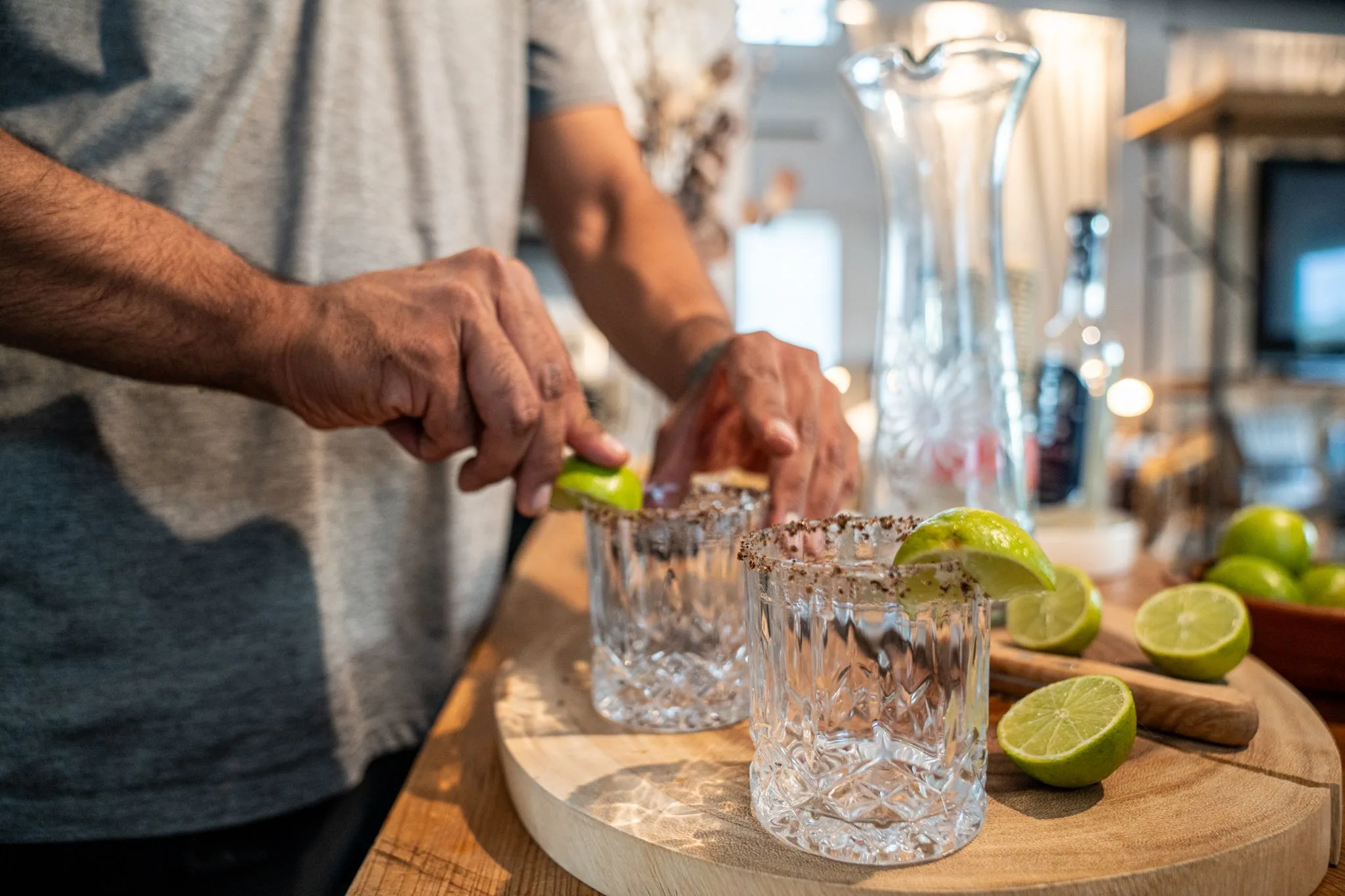 Hands squeezing limes over two salt-rimmed crystal glasses on a wood board
