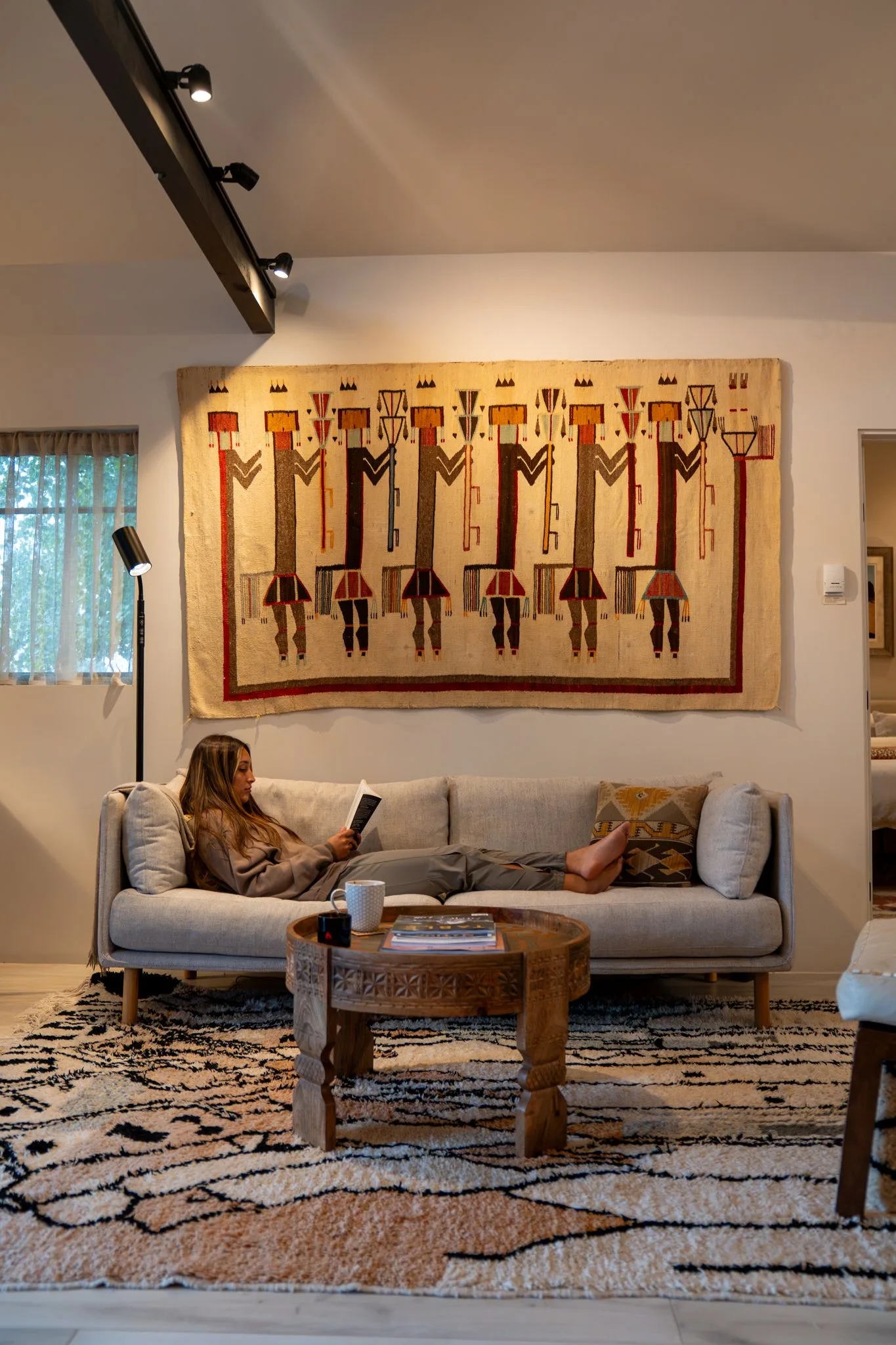 A guest reading on the gray sofa in the Farmhouse 2 living room, beneath a large Navajo Yei weaving, lamplight on the white walls