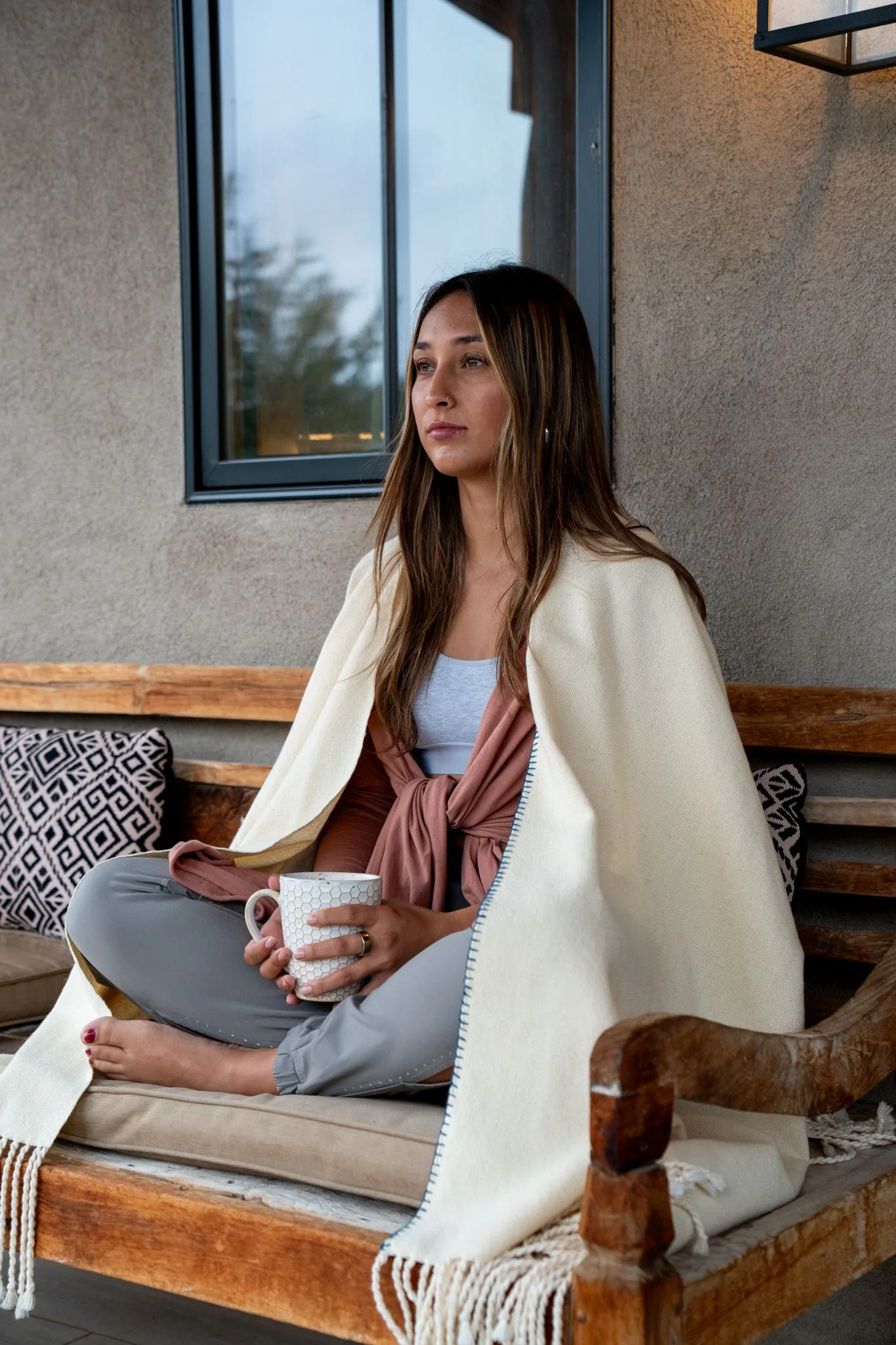 A woman wrapped in a cream blanket sits cross-legged on a wooden bench on the Farmhouse 2 porch, holding a white mug in the morning light
