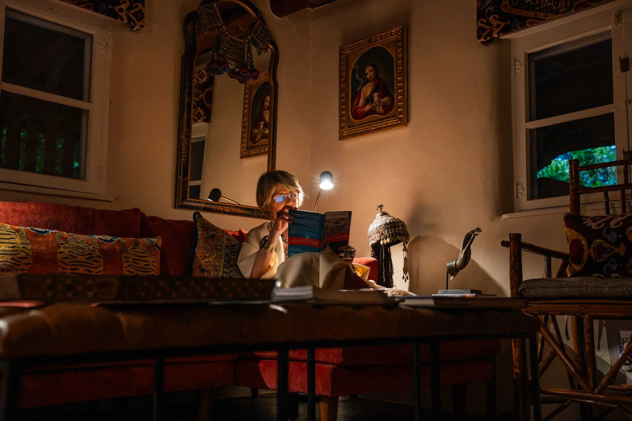 A woman reads by a single lamp on the red sofa in the Los Santos sitting room, candles and carved mirrors around her