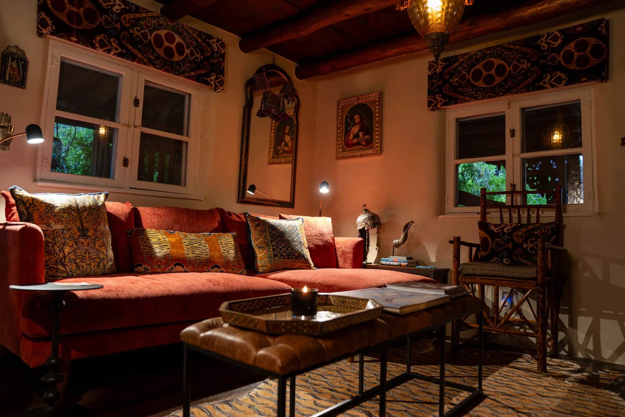 A sitting room at Los Santos with a red sofa, embroidered pillows, carved mirrors, and an ottoman under lamplight