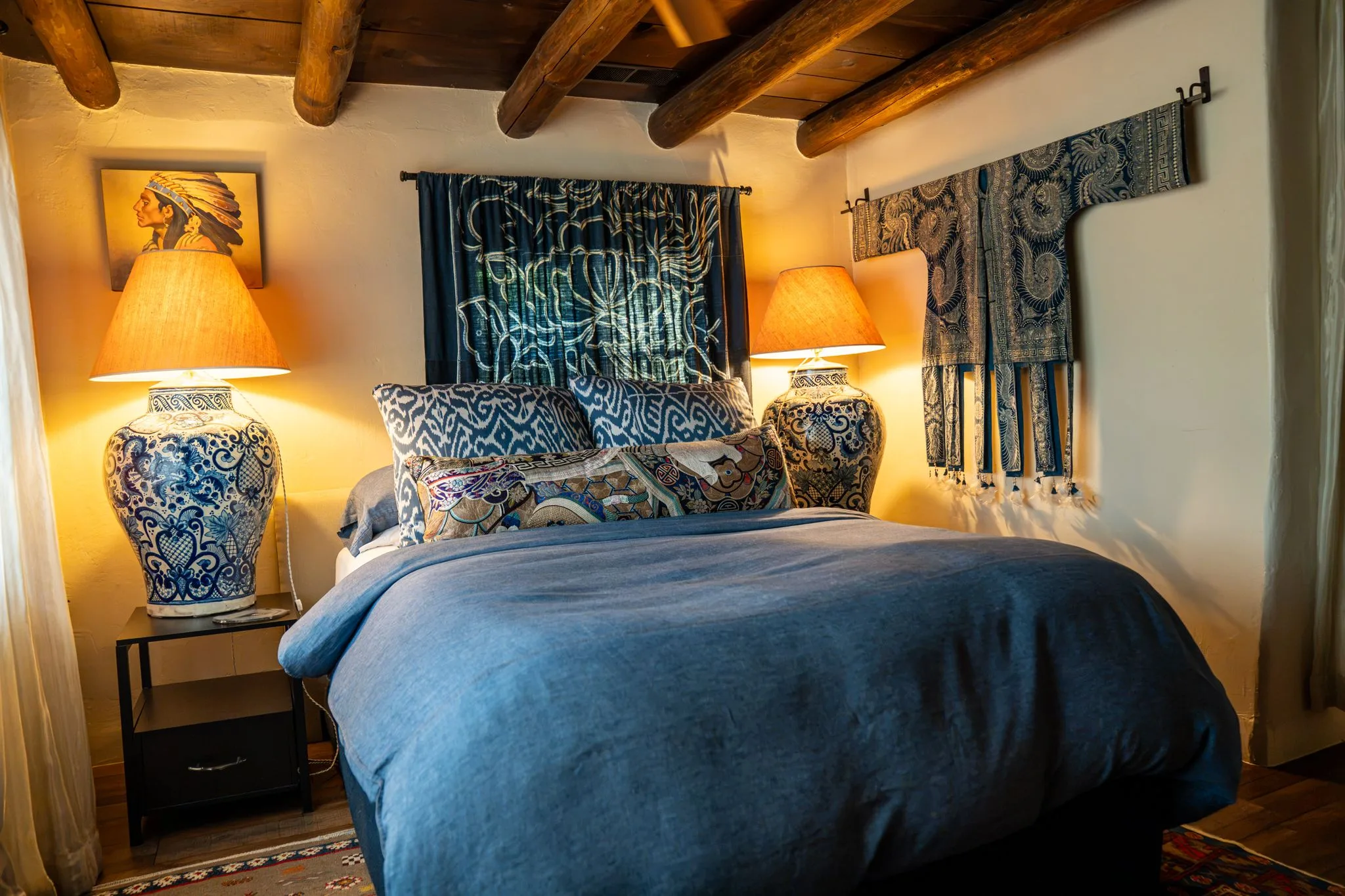 A bedroom with a dark blue duvet, blue and white ginger jar lamps, and an indigo textile hung over the bed under vigas