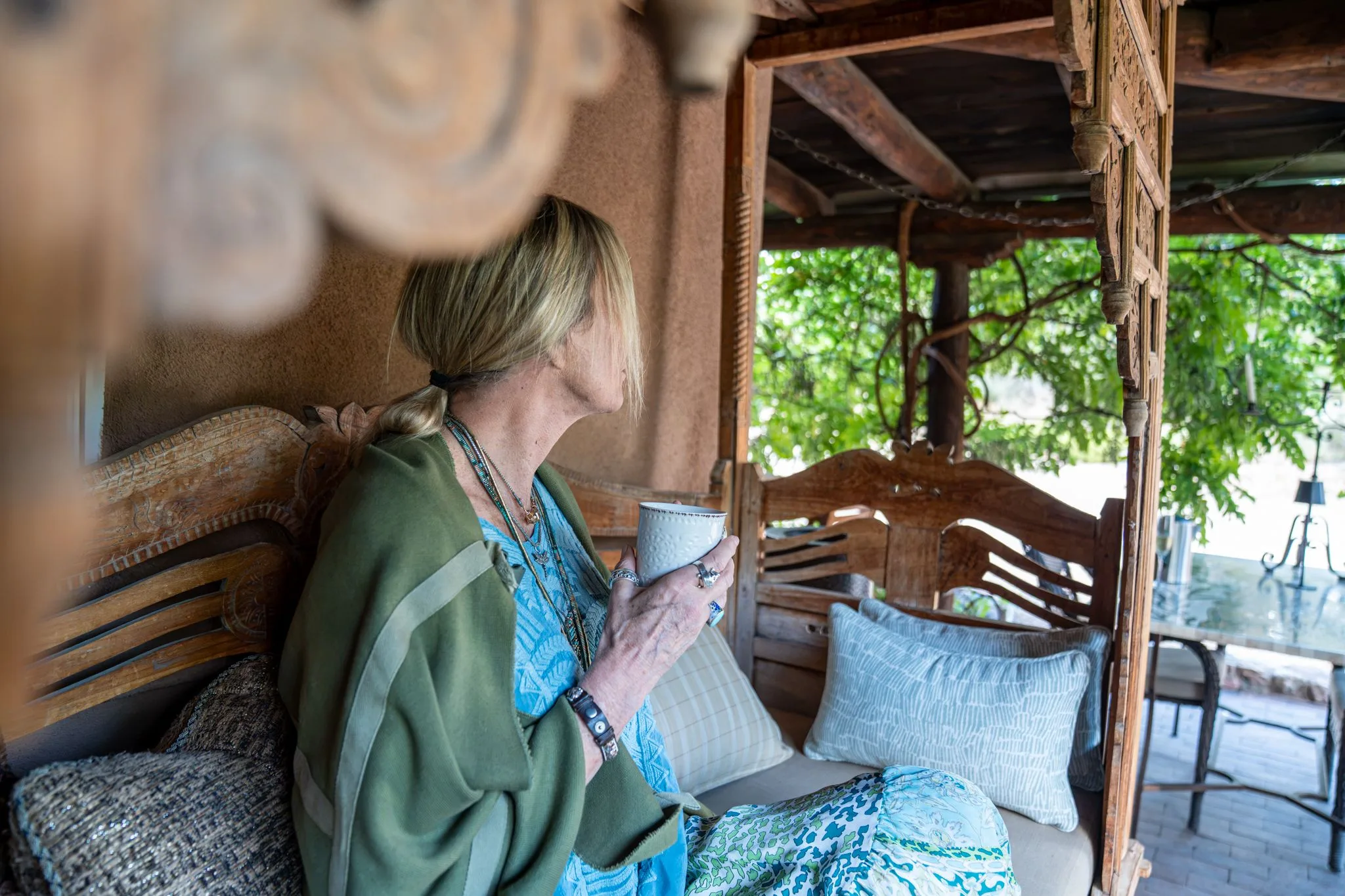 A woman in a green wrap holds a cup on the carved daybed of the Los Santos portale, the garden beyond