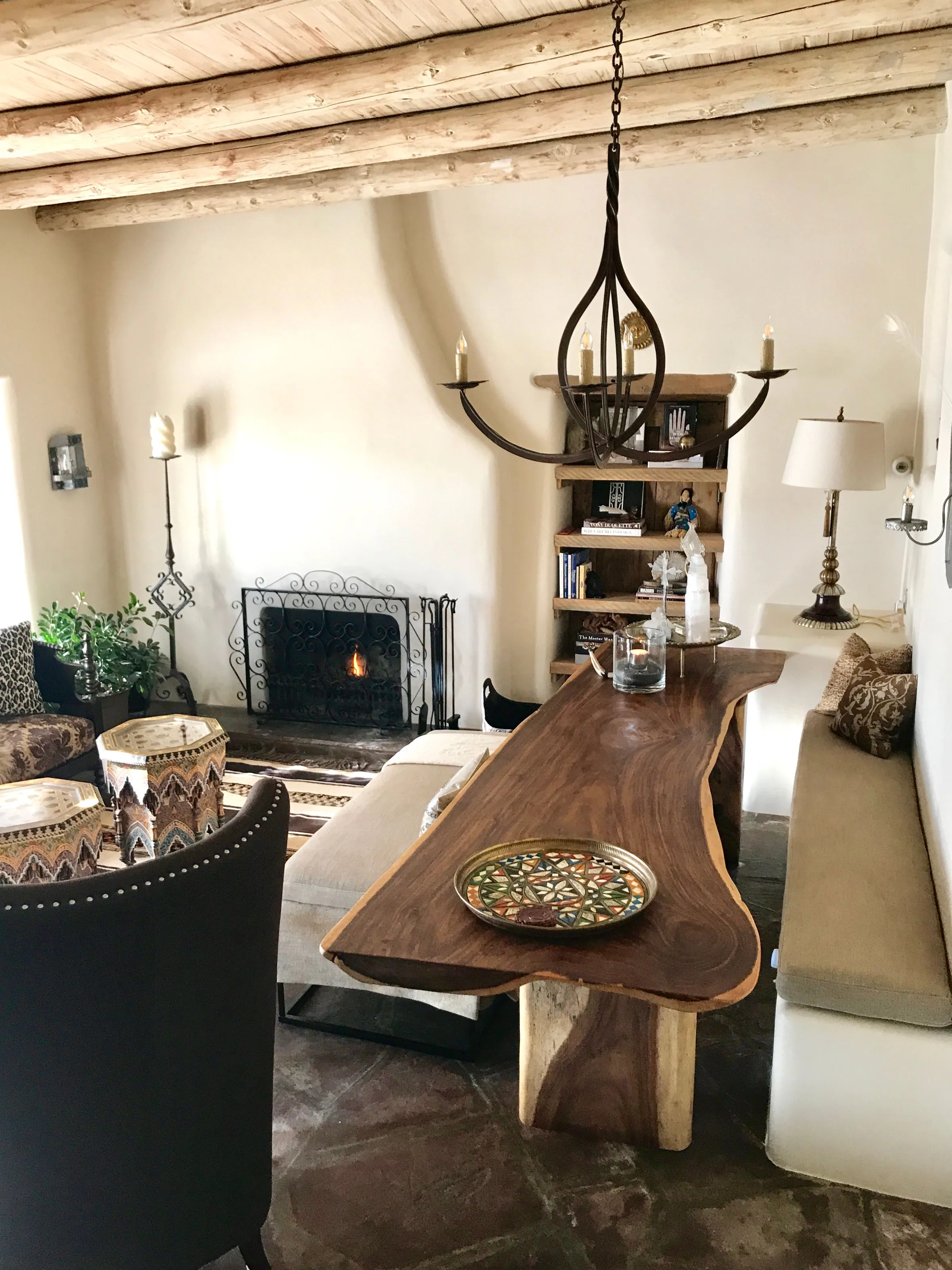 The Los Santos sala with a fire burning behind an iron screen, a live-edge nine-foot dining table, a banco with cushions, and bookshelves set into the adobe wall