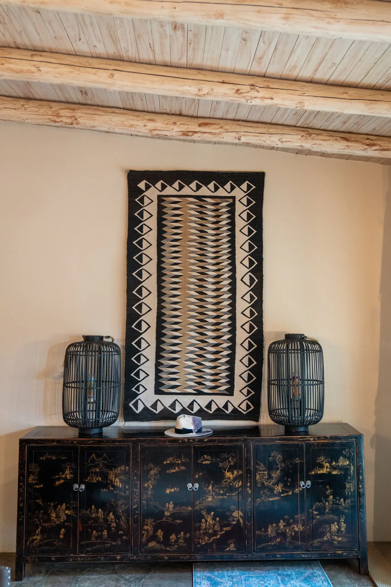 A carved black chinoiserie chest with two woven lanterns beneath a framed Navajo weaving under pale vigas