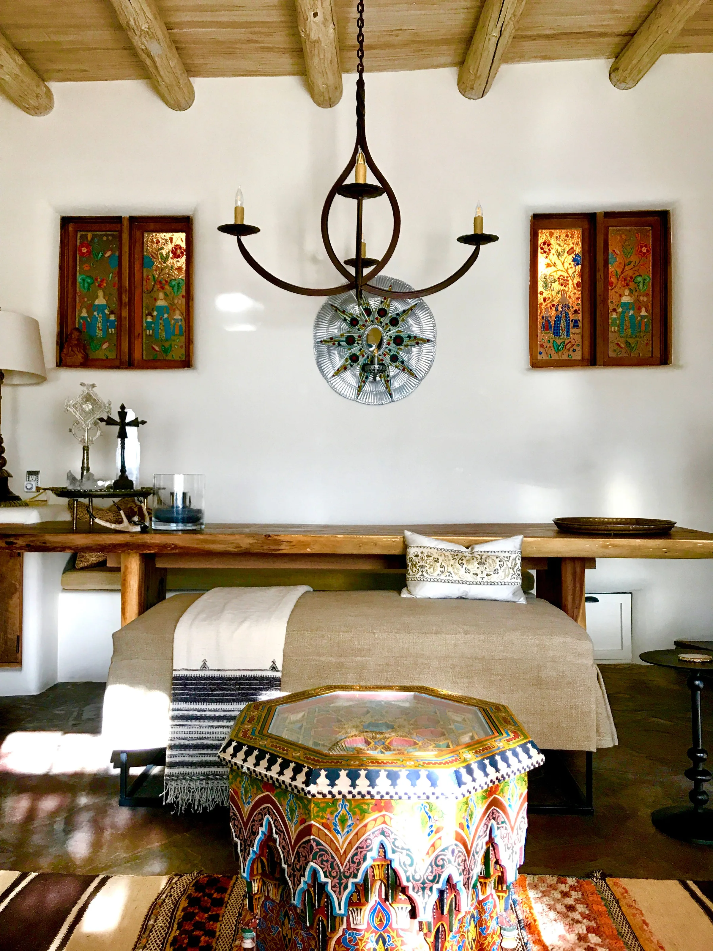 The Los Santos sala: a long wooden table and a daybed under the vigas, an iron chandelier, painted shutters on white walls, and a beaded ottoman