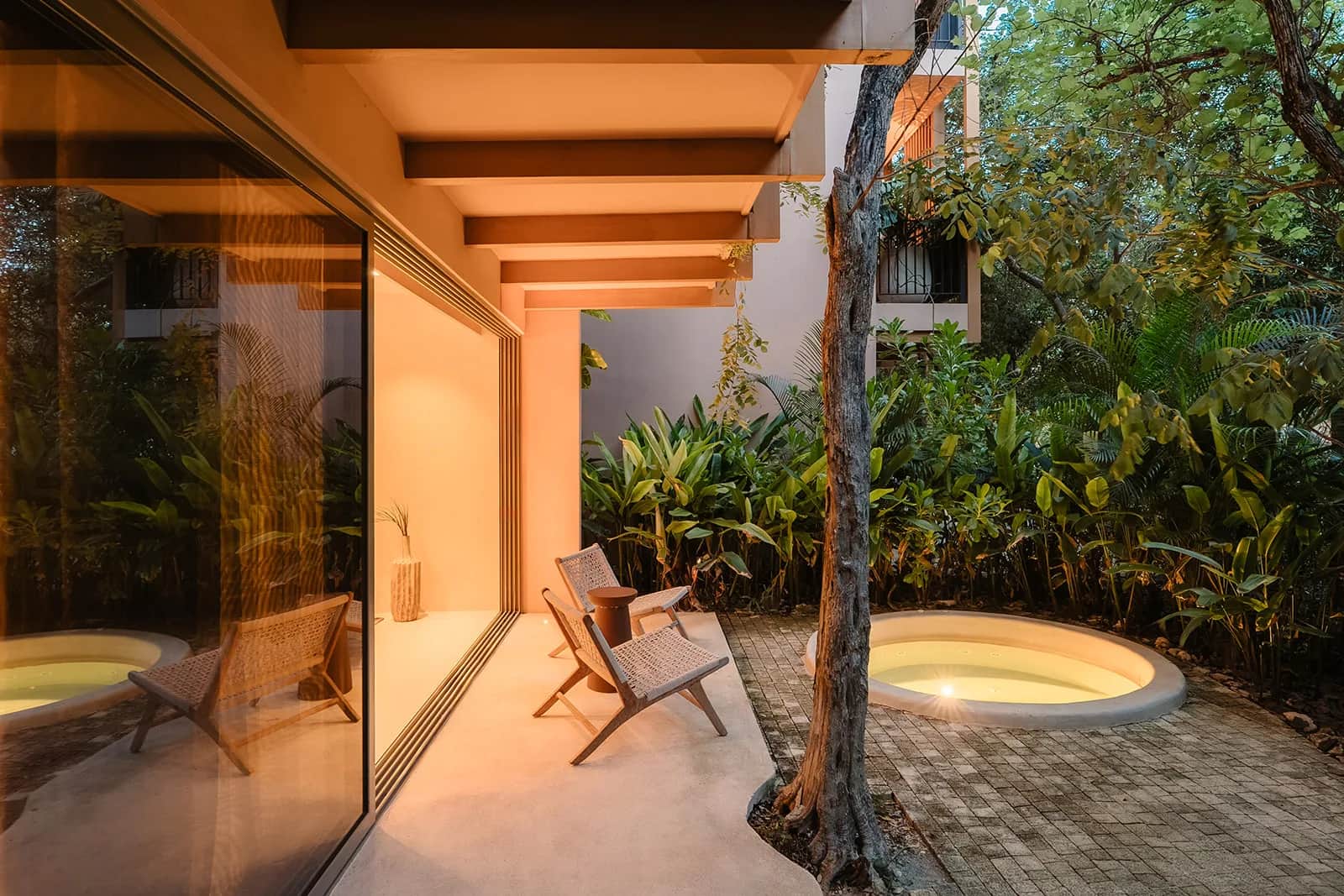 A private courtyard at dusk with a round jacuzzi, two woven chairs, and jungle trees beside the glass walls of the apartment