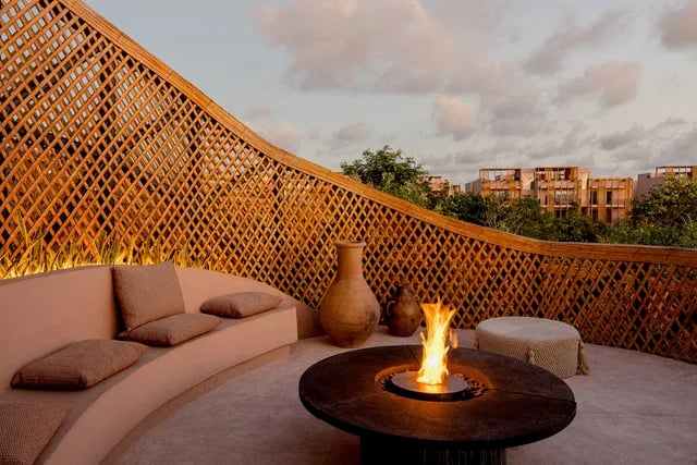 A rooftop lounge at dusk: a curved bench, a fire bowl, and a lattice wall looking over the treetops