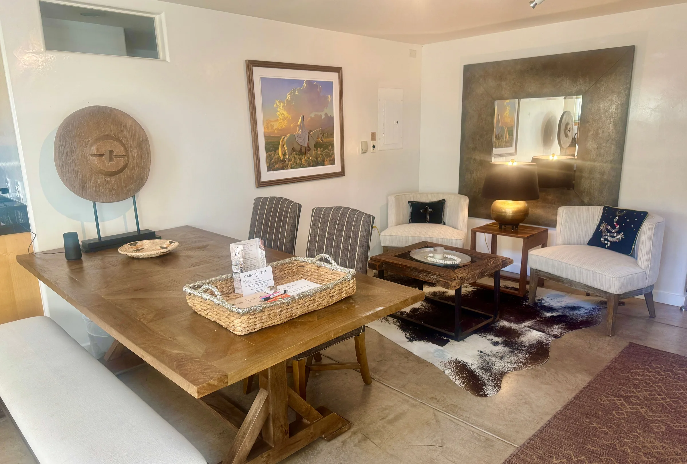 The dining table set with a woven tray, white armchairs and a dark coffee table beyond, a painting of horses and a bronze mirror